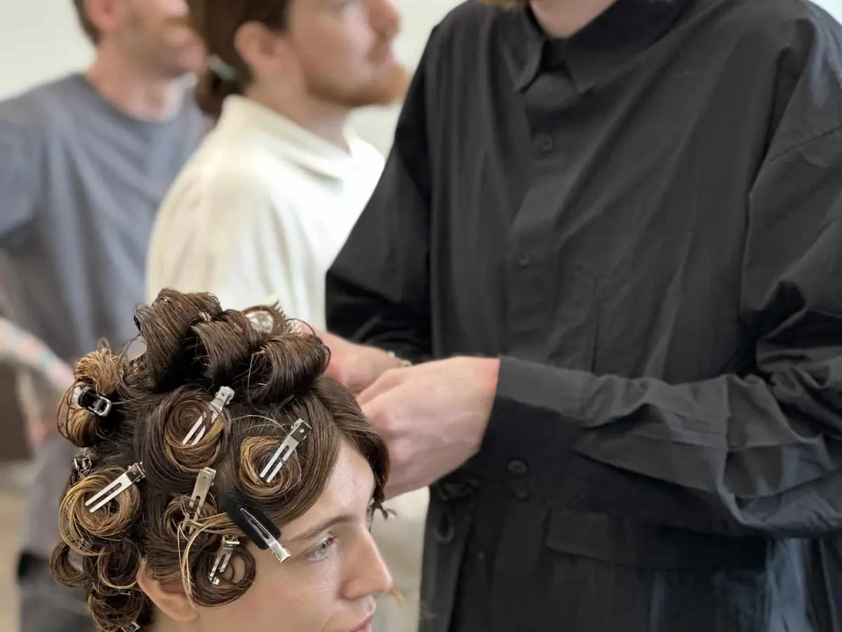 A cosmetology instructor in black works on a seated student's hair set in rollers while other people practice in the classroom behind them.