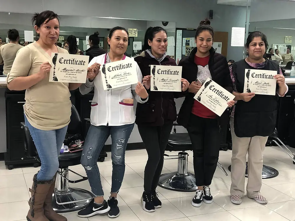 Five women stand inside a salon classroom with styling chairs and mirrors, each holding a certificate.