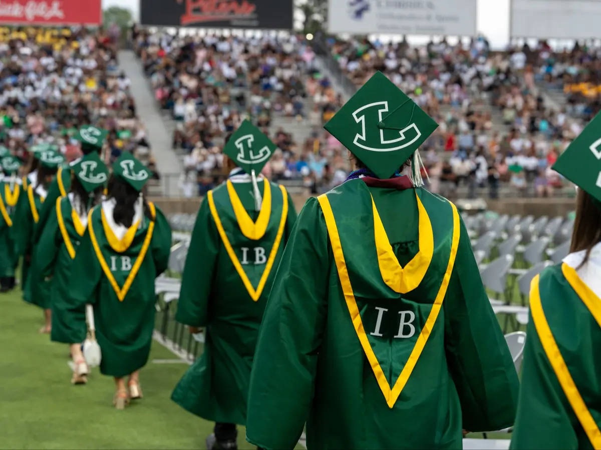 Longview High School students in green graduation robes with gold trim and L caps walk across a football field during an IB ceremony, with a crowded stadium in the background.