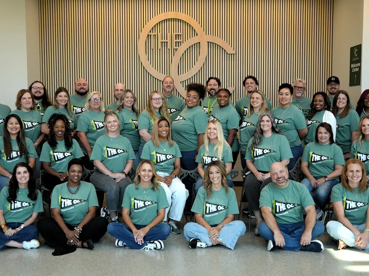 A large group of staff members wearing teal The OC shirts pose indoors beneath a large circular OC logo on a wood-slat wall.