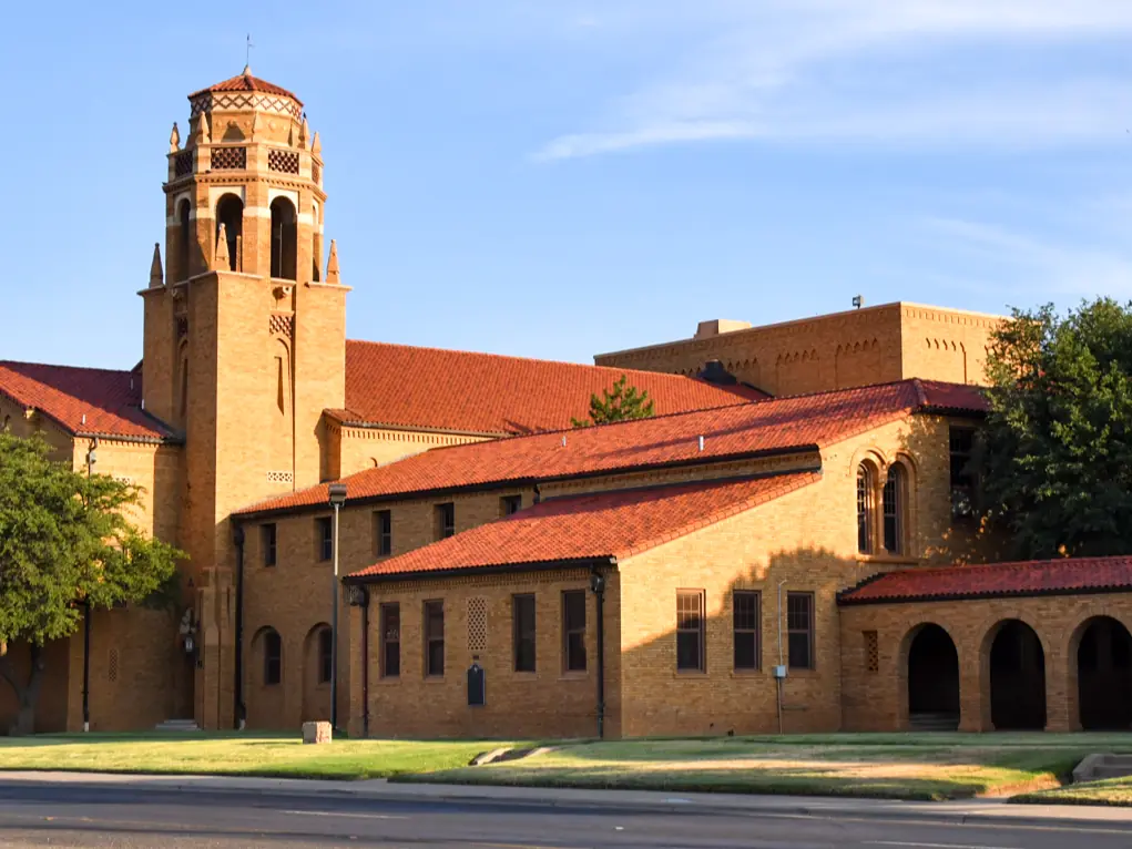 A tan-brick school building with red tile roofs, arched walkways, and a tall decorative tower under a blue sky.