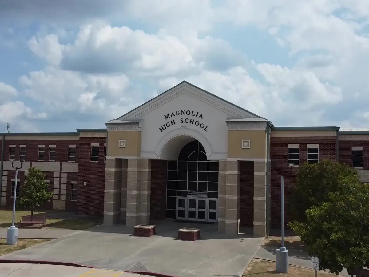 Front entrance of Magnolia High School, a broad red-brick school building with a central stone facade, arched glass doorway, and the school name displayed above it under a partly cloudy sky.