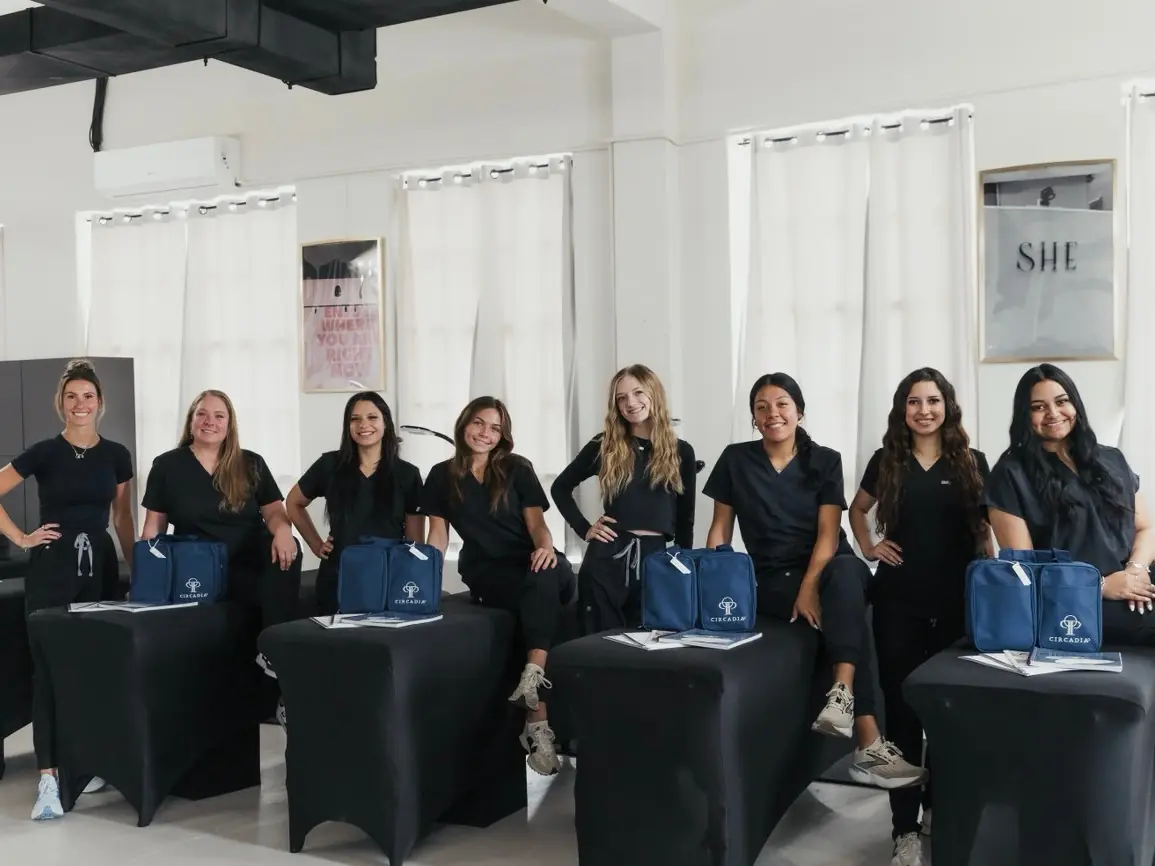 Seven women in black clothing stand and sit among black salon styling stations in a bright room with white curtains and blue bags.