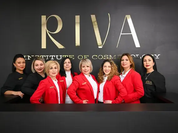 Seven Riva Institute of Cosmetology staff members stand behind a counter beneath a large RIVA Institute of Cosmetology logo on a dark wall.