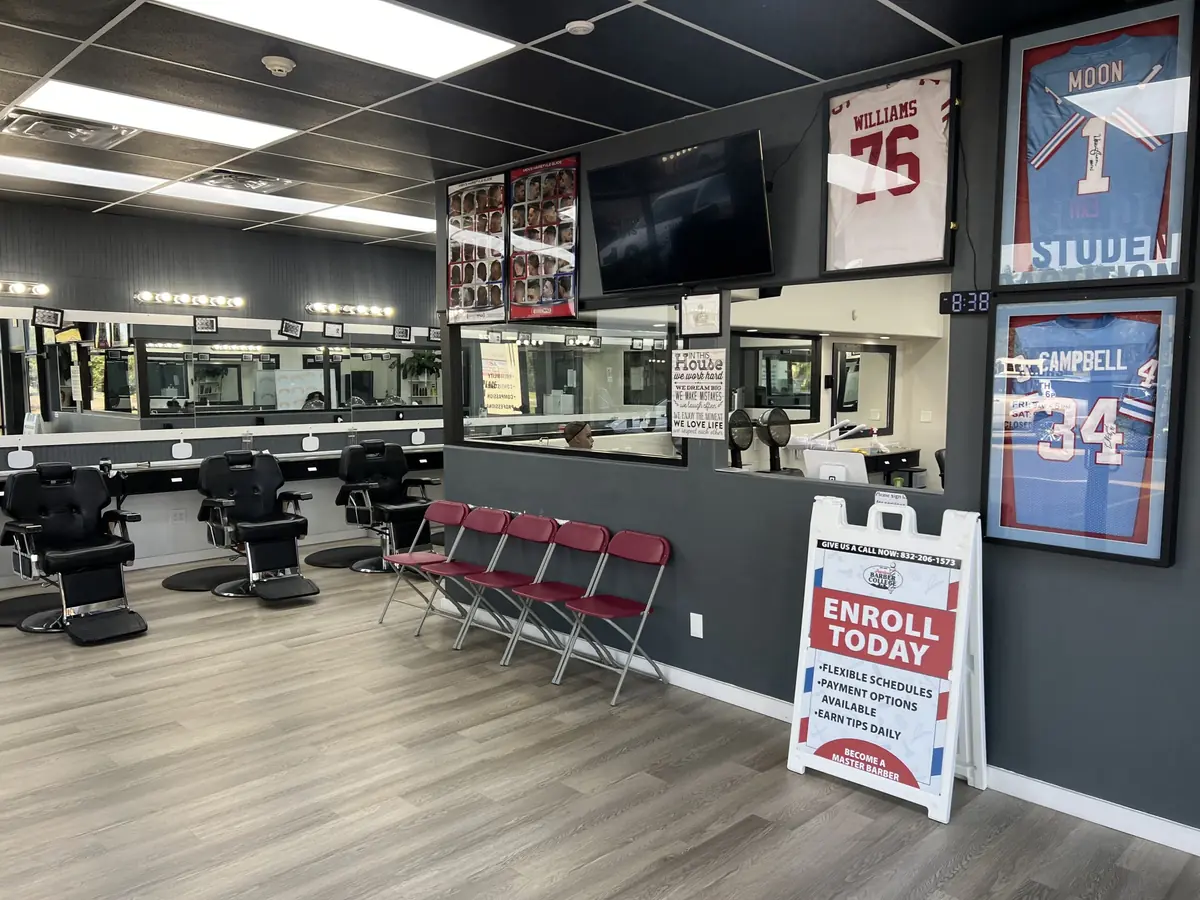 Interior of Pearland Barber College with rows of black barber chairs and mirrors, a wall-mounted television, framed sports jerseys, and an A-frame sign reading “ENROLL TODAY.”