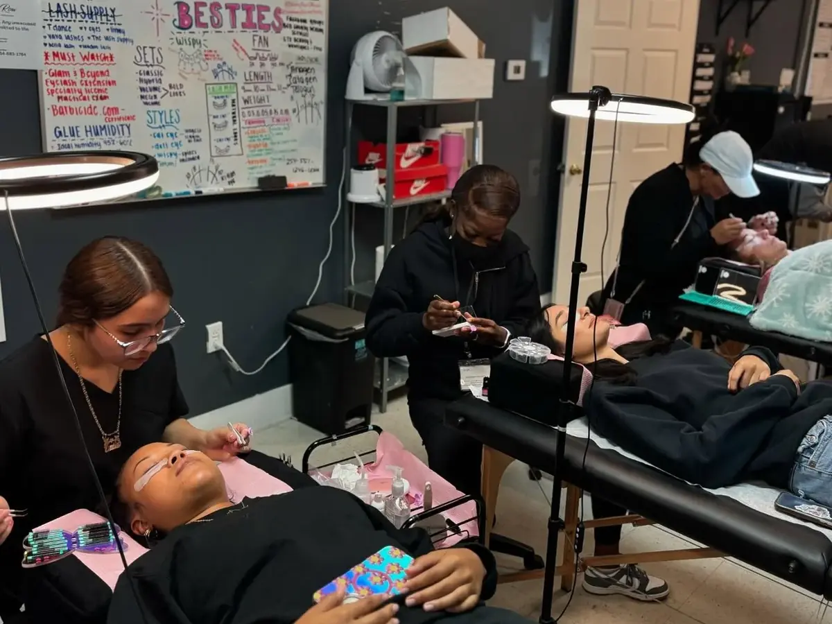 A beauty-training classroom with several students practicing services on clients at salon beds and tables, ring lights, and a wall covered with instructional notes.