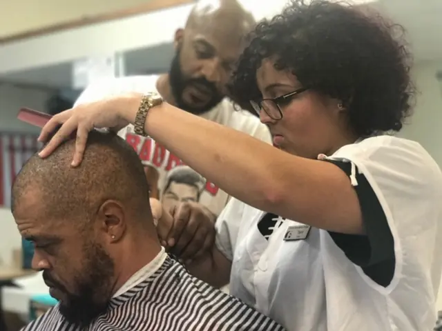 A barber instructor in a white uniform trims a seated man's hair while another person observes in an indoor barber-training area.