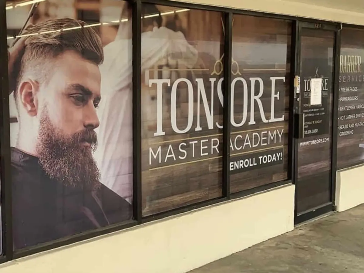 Exterior storefront of Tonsore Master Academy with large window graphics showing barbering images and the words “TONSORE MASTER ACADEMY” and “ENROLL TODAY.”