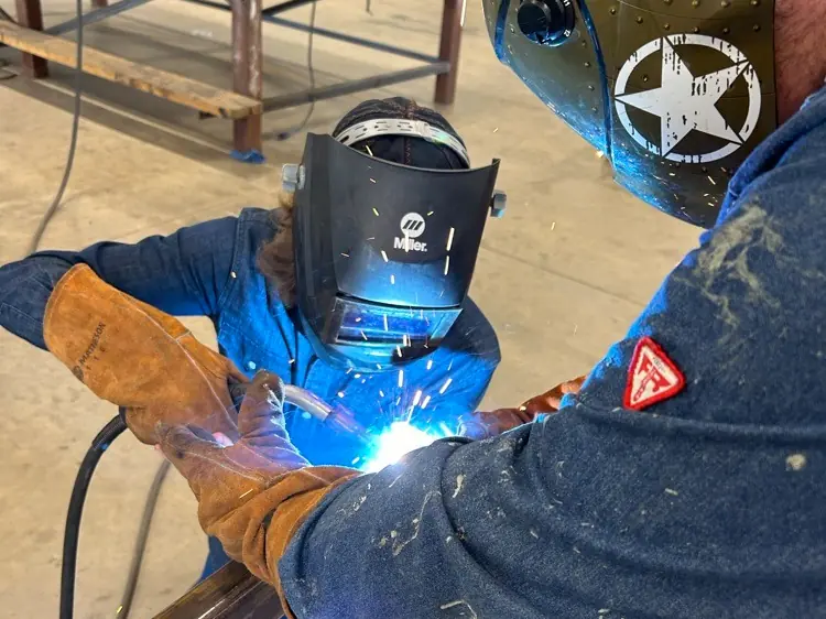 Two students wearing welding helmets and protective gloves weld metal at worktables in a school welding classroom.