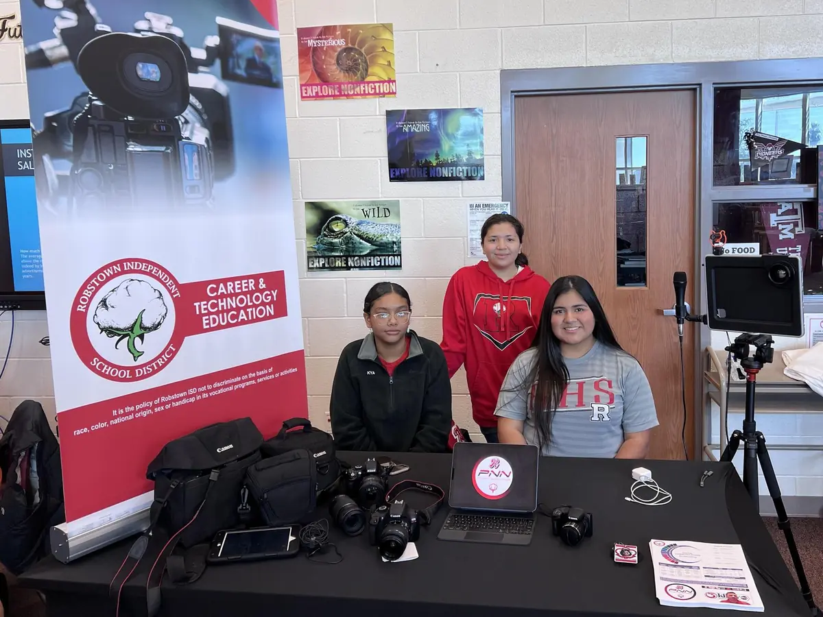 Three students stand behind a table displaying cameras, video equipment, and a laptop in a Robstown ISD Career and Technology Education media-production space; a fourth person is partially visible at the left.