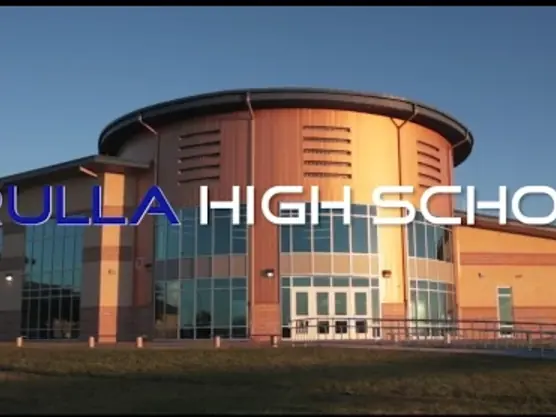 Exterior photograph of the Grulla High School building, showing a modern curved orange-and-glass entrance under a blue sky.