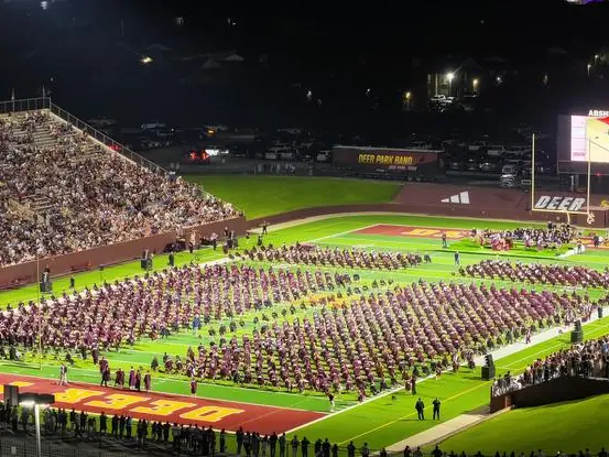A nighttime graduation ceremony fills the football field at Deer Park’s stadium, with rows of graduates on the field, a large crowd in the stands, and a lit stage and video board.