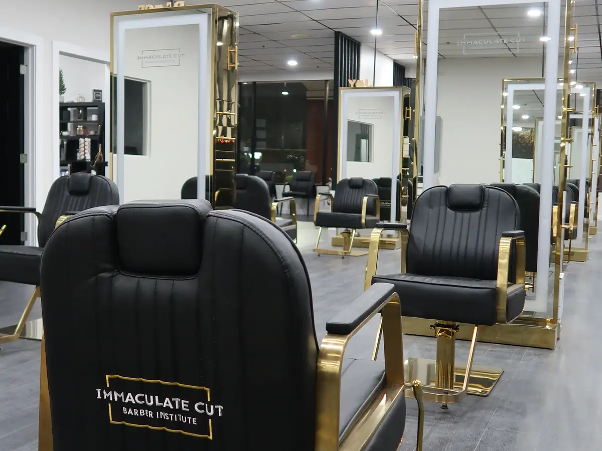 Interior of a barber institute with black and gold barber chairs, mirrors, and bright ceiling lights; the nearest chair is branded Immaculate Cut Barber Institute.
