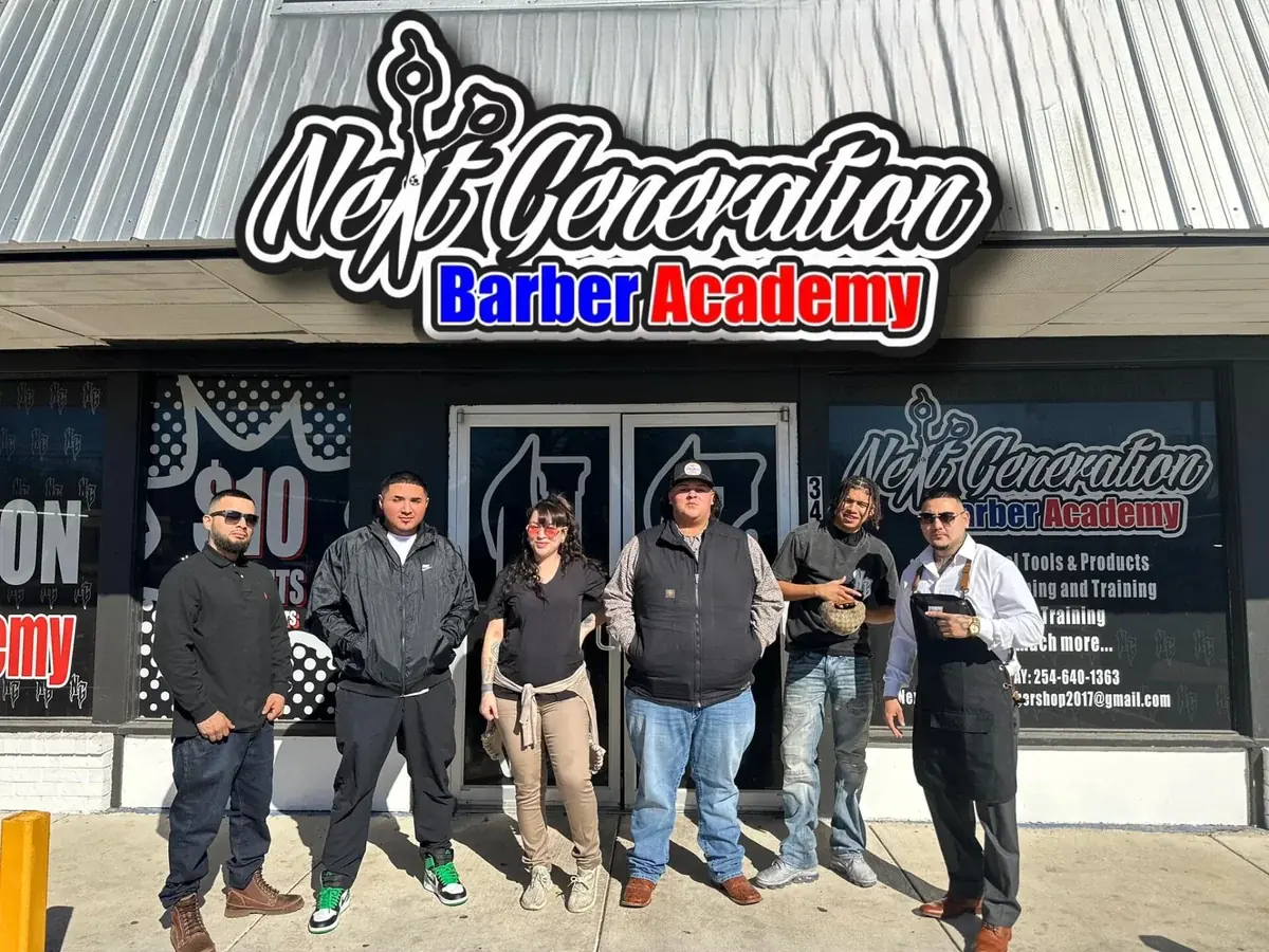 Six people stand outside the Next Generation Barber Academy storefront beneath a large academy sign; the building has barber academy graphics on the windows and a glass entrance.
