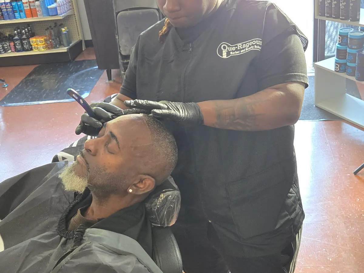 A barber wearing a Querageous Barber School shirt trims a seated client's hair in a salon, with product shelves and barber stations visible around them.