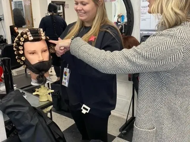 Two students practice hairstyling on a mannequin in a salon classroom, with mirrors, shelves, and styling equipment visible.