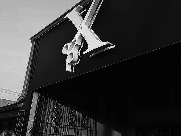 Black-and-white exterior view of the Xotic Barber Academy storefront, with a large scissors emblem mounted above the entrance and barred windows along the facade.
