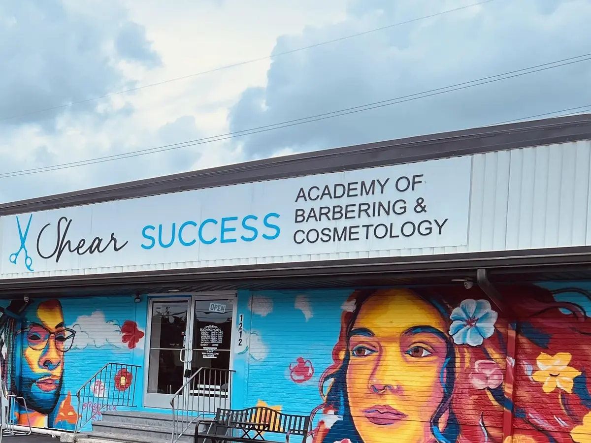 Exterior of Shear Success Academy of Barbering & Cosmetology in a one-story building with a colorful mural, a bench by the entrance, and marked parking spaces in front.