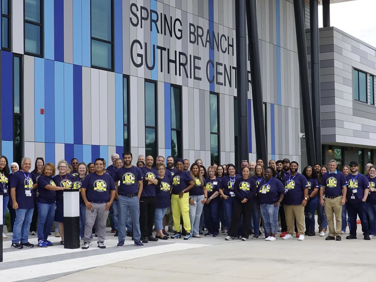 A large group of people in matching blue and purple shirts stands outside the modern Spring Branch Guthrie Center building, whose facade displays the words “SPRING BRANCH GUTHRIE CENTER.”