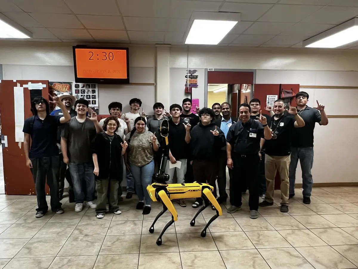 A group of students and adults pose in a tiled school hallway behind a yellow wheeled device, with an orange digital clock and classroom doors in the background.