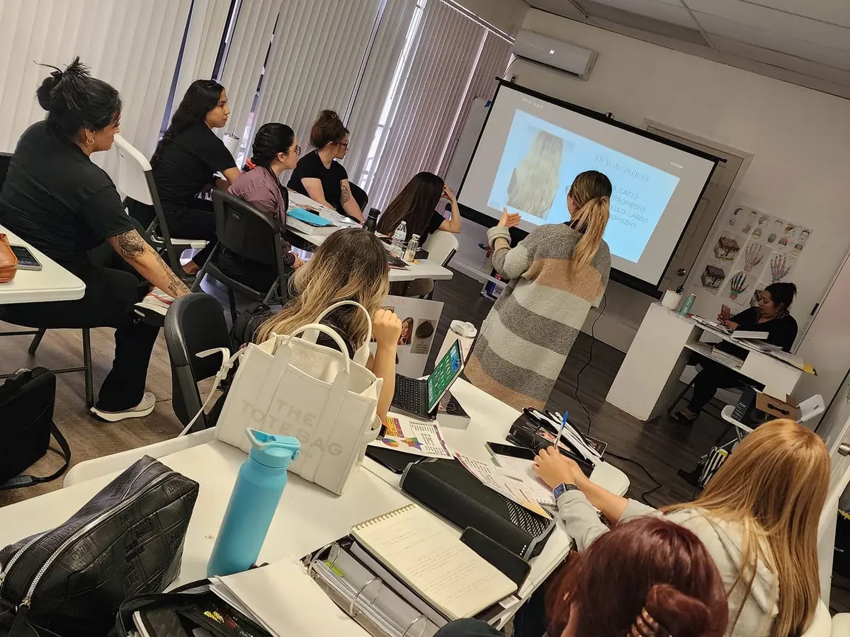 Students seated at tables in a beauty academy classroom while an instructor presents material on a projection screen.