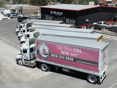 Aerial view of a Prime Time CDL training facility with a black building bearing the school sign, several white tractor-trailers parked in rows, and a large box truck in the foreground.
