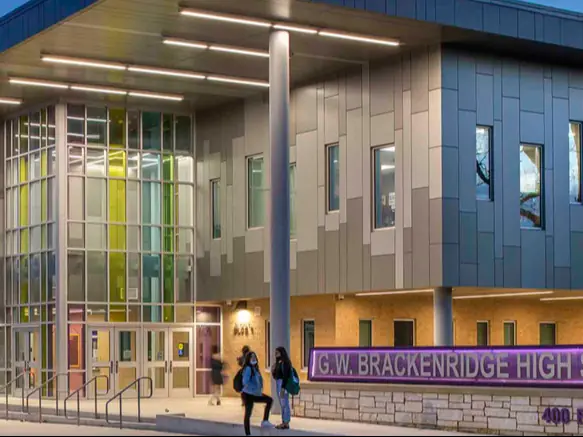 Exterior of G. W. Brackenridge High School, a modern gray building with a glass entrance, blue roofline, and purple school sign beside the walkway.