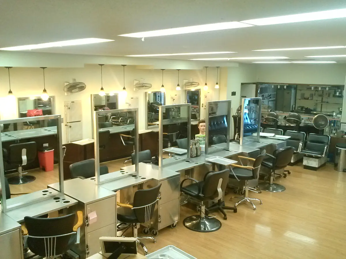 Cosmetology training salon with rows of styling stations, mirrors, salon chairs, hood dryers, and work counters under fluorescent ceiling lights.