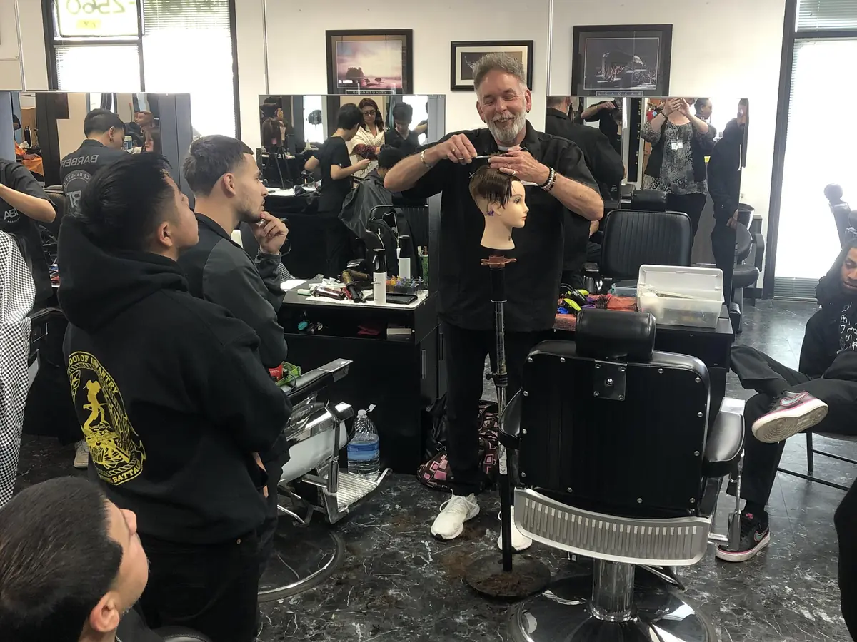 Barbering students gather around an instructor demonstrating a haircut on a mannequin in a salon classroom, with mirrors, styling stations, and chairs visible throughout the room.