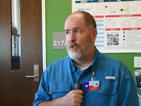 A man in a blue work shirt stands inside a room beside a door marked 217; a calendar and QR code are posted on the wall behind him.