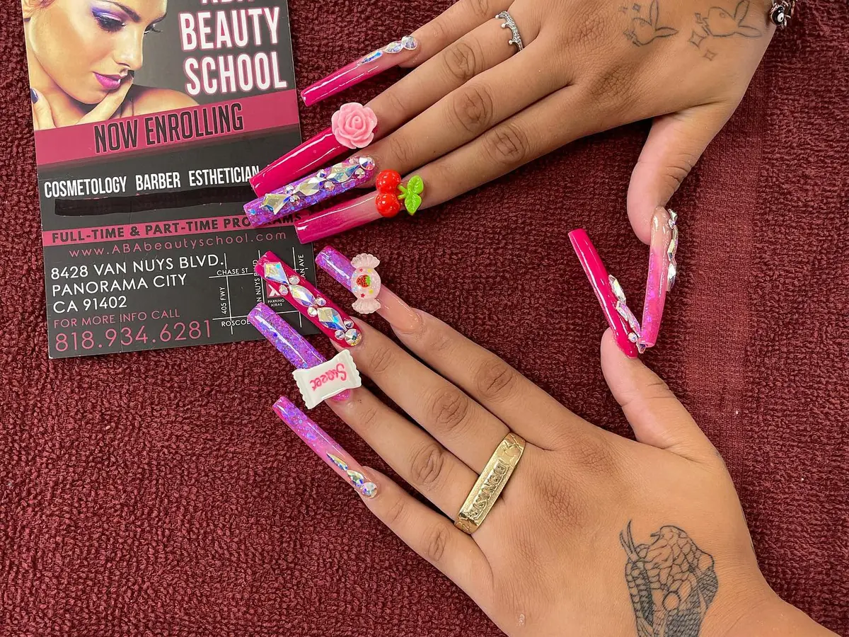 Two tattooed hands display long, colorful decorated acrylic nails over a burgundy towel beside an American Beauty School enrollment card listing the Panorama City address.