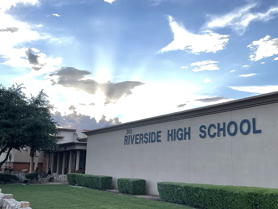 Exterior of Riverside High School building with the school name displayed on the facade, trees and landscaped grounds in front, and a partly cloudy sky above.