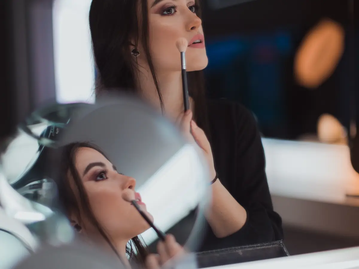 Student applying makeup to a seated client at a salon mirror, with additional reflected faces and makeup brushes visible in the foreground.
