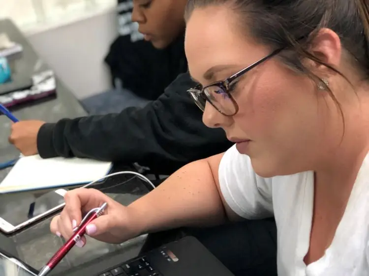 Two students work at laptops and write notes at a table in a beauty-school classroom.