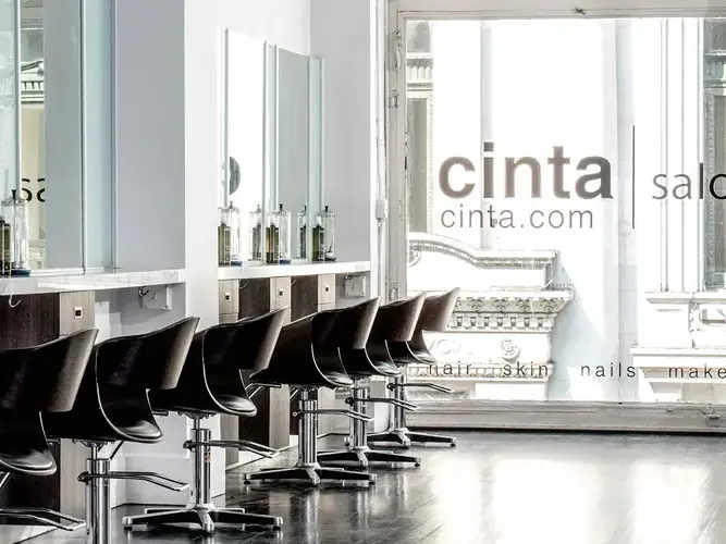 Bright Cinta Aveda salon interior with rows of black styling chairs, mirrors, sinks, product shelves, and large windows.