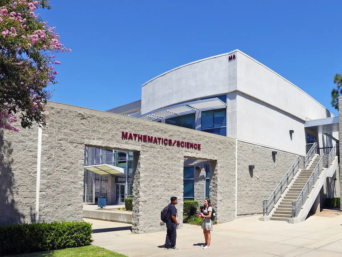Exterior of Citrus College’s Mathematics/Science Building, a low gray-stone and white structure with red lettering, large windows, and an outdoor staircase under a clear blue sky.