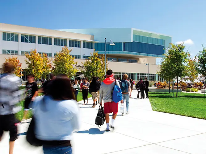 Students walking along a campus pathway toward a large modern College of San Mateo building, with a broad green lawn and trees in front.
