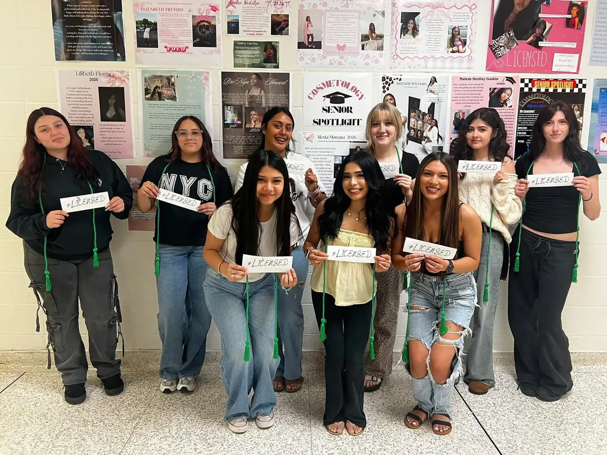 A group of nine students pose indoors in front of a cosmetology student spotlight display, holding small cards that say “LICENSED.”