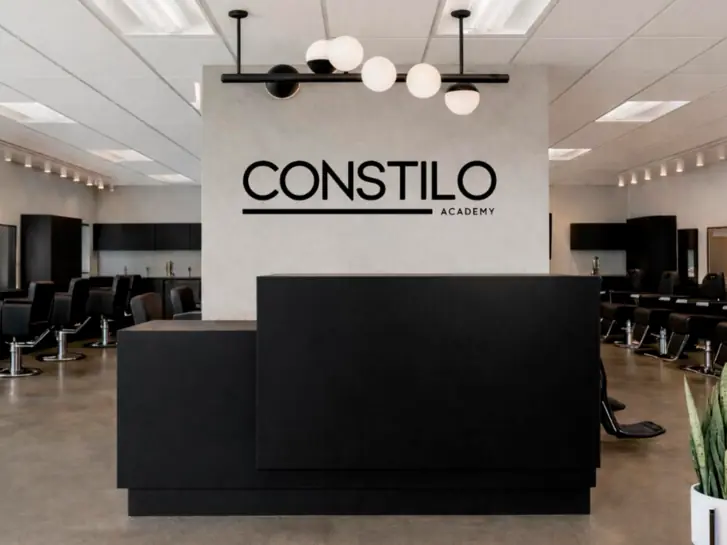 Interior of Constilo Academy showing a black reception desk, the CONSTILO ACADEMY logo on the wall, salon stations lining both sides of the room, ceiling lights, and a potted plant in the foreground.