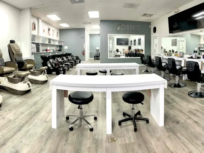 Bright Cosmo Beauty College student salon with manicure tables in the center, pedicure chairs along the left wall, styling stations and mirrors along the right wall, and the Cosmo logo above the rear doorway.