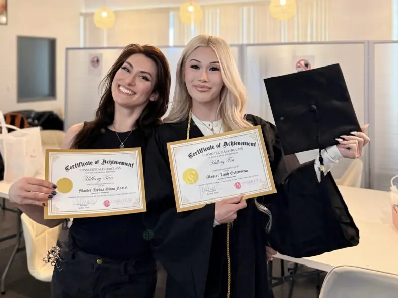 Two smiling Cosmotek College students hold Certificates of Achievement in a bright salon-style training room with pendant lights, mirrors, and salon chairs.