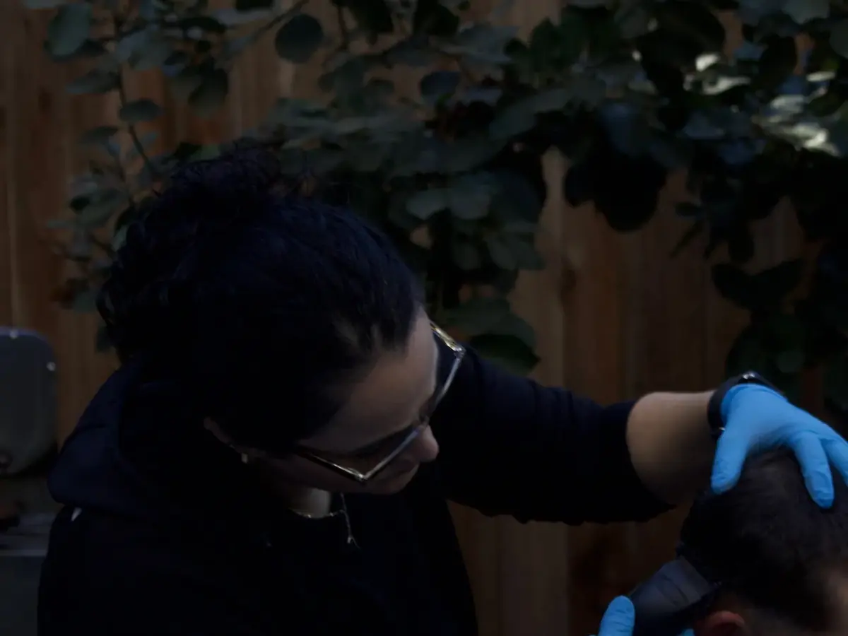 A barber wearing blue gloves trims a seated client's hair with clippers in a cosmetology service area.