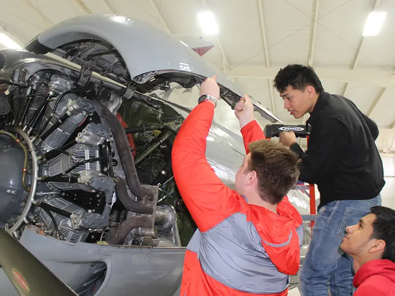 Three students work around an exposed aircraft engine inside a brightly lit workshop; one student in a red-and-gray jacket reaches up to the engine while two others observe.