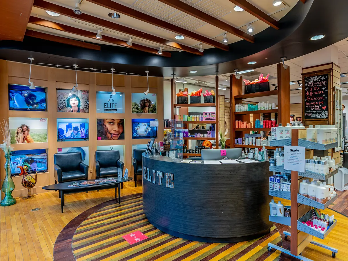 Interior of Elite Cosmetology School’s student salon with a round ELITE reception desk, salon chairs, product shelves, wall-mounted screens, and wood flooring.