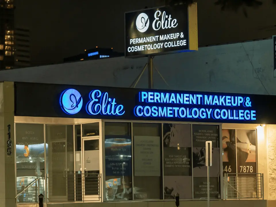 Nighttime exterior of Elite Permanent Makeup & Cosmetology College, with illuminated blue storefront signage, glass entrance, and the college sign mounted above the building.