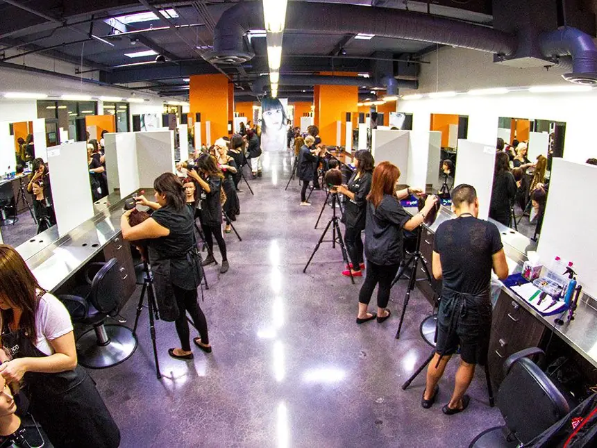 Wide interior view of a busy beauty school salon with students working at rows of styling stations and mirrors.