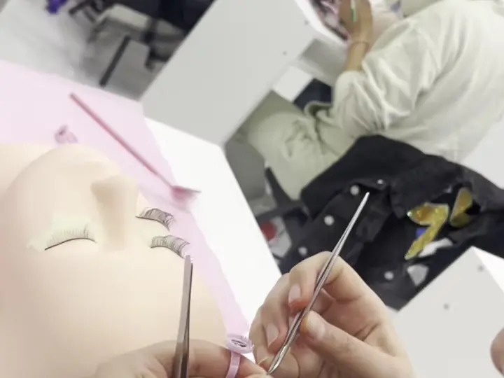 Close-up of a student using tweezers on a practice lash or brow treatment while other students work at salon stations in the background.