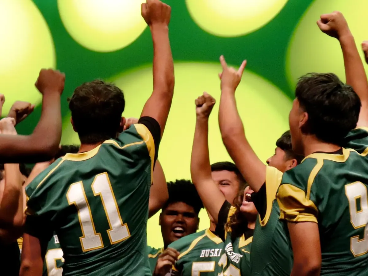 Holmes High School students in green football uniforms celebrating together with their arms raised.