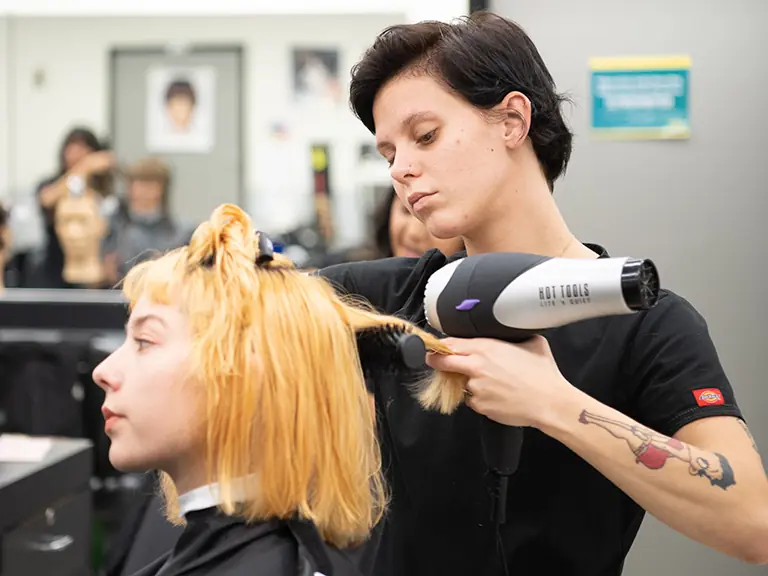 A cosmetology student uses a hair dryer and round brush to style a client's blonde hair in a salon classroom, with mirrors and other workstations behind them.