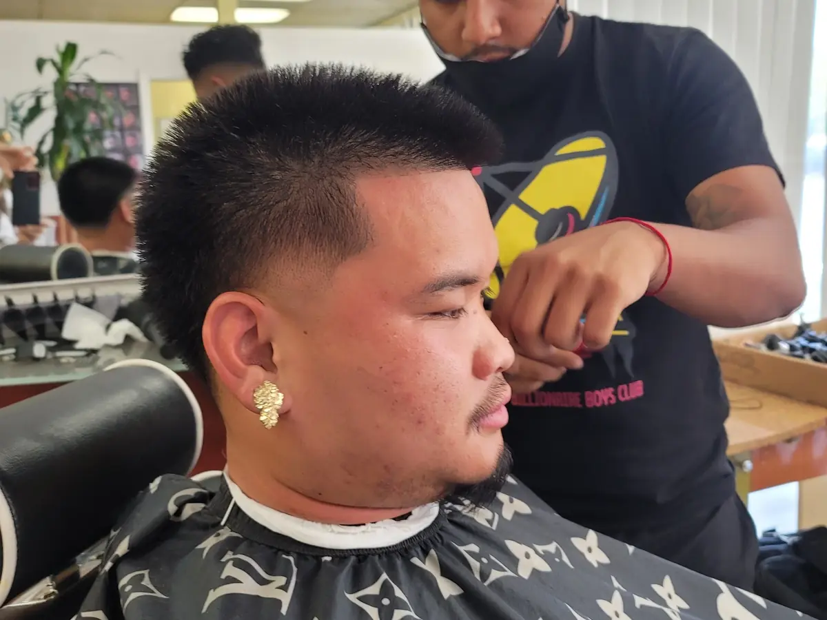 A barber trims a seated client's hair inside a bright cosmetology classroom, with salon stations and mirrors visible behind them.